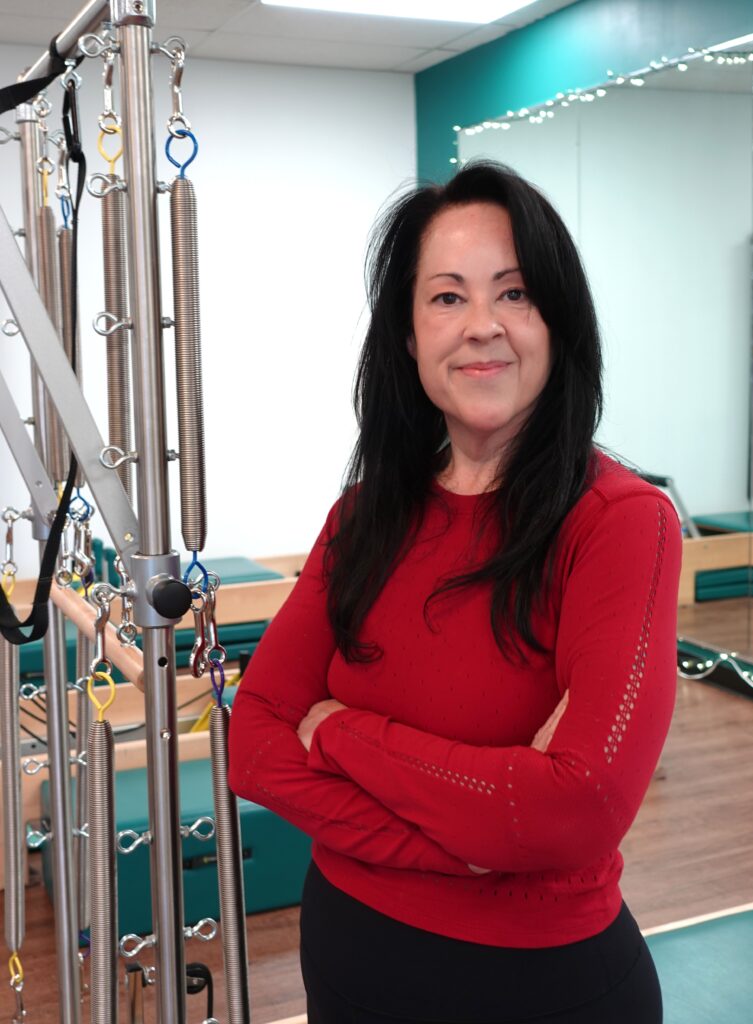 Headshot of instructor Sandra Rocha. Standing in front of the Pilates equipment in the studio, smiling not showing teeth, long dark brown hair, wearing a red long sleeve top.