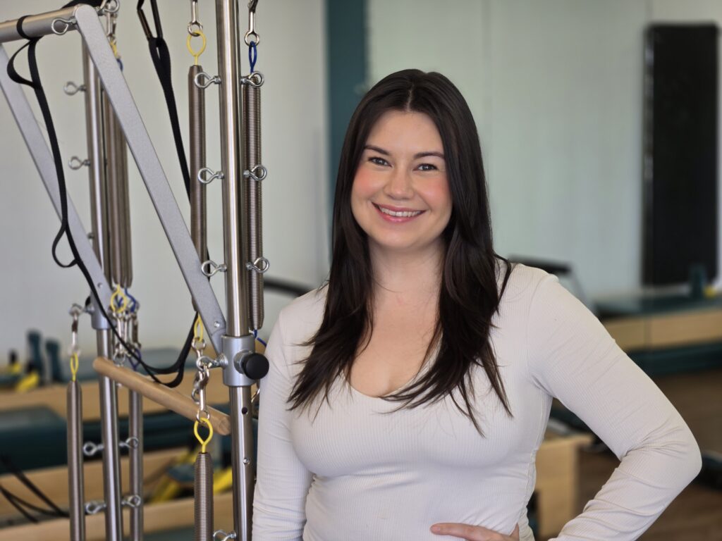 Headshot of Taylour. Standing in front the of the Pilates equipment. Medium length dark brown hair worn down with a white long sleeve top