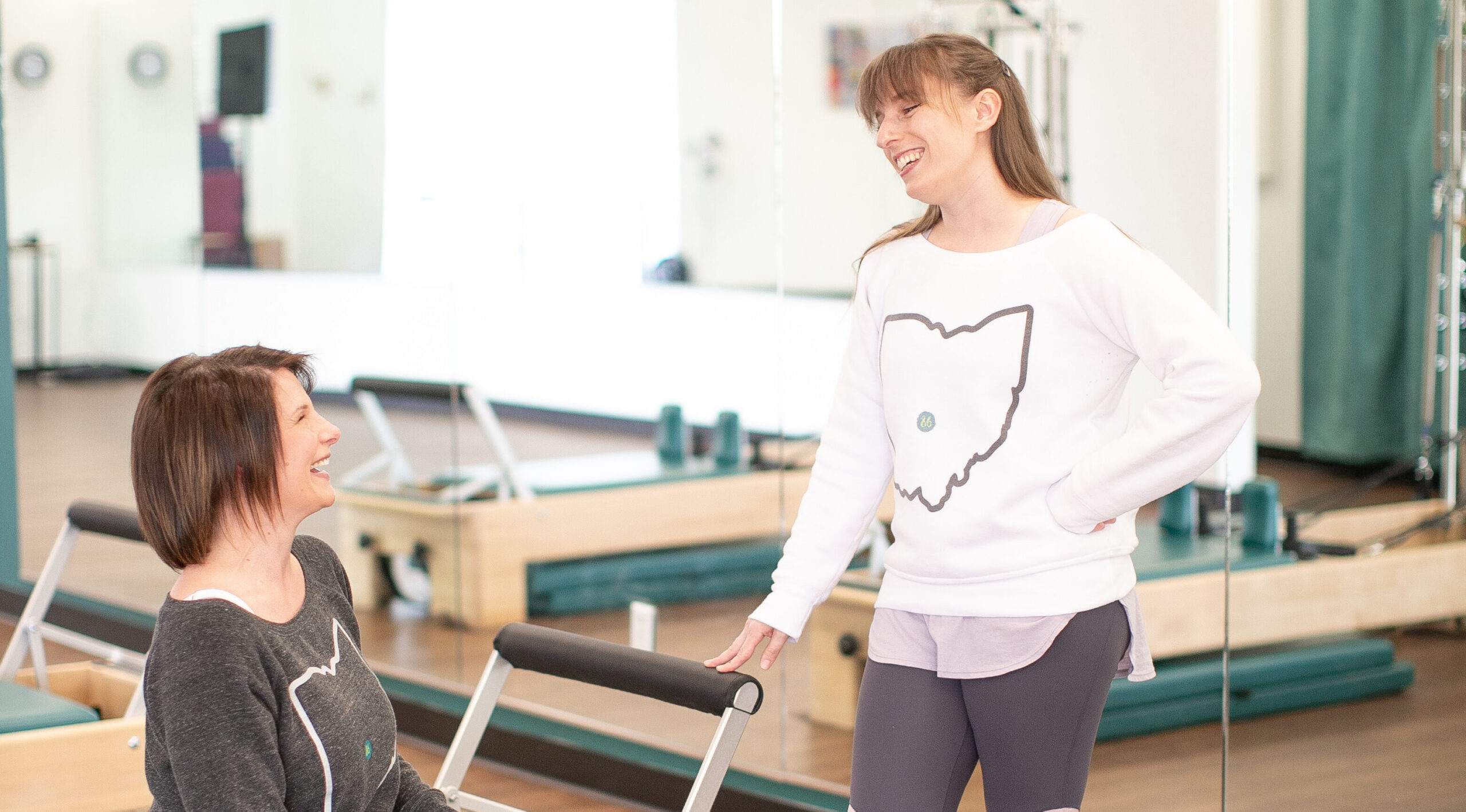 Two women talking while standing near a Pilates Reformer. Both women are smiling.